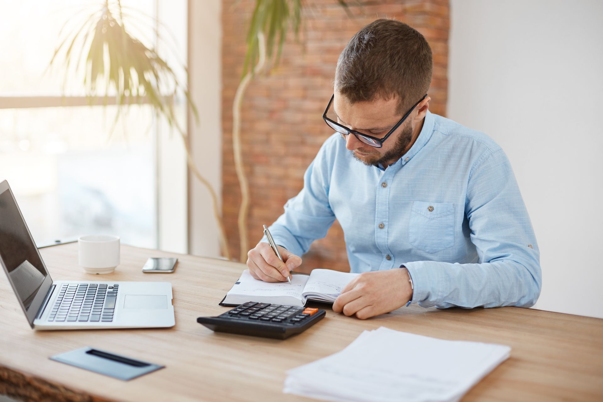 Young man with glasses calculating expenses with a calculator and writing in a notebook