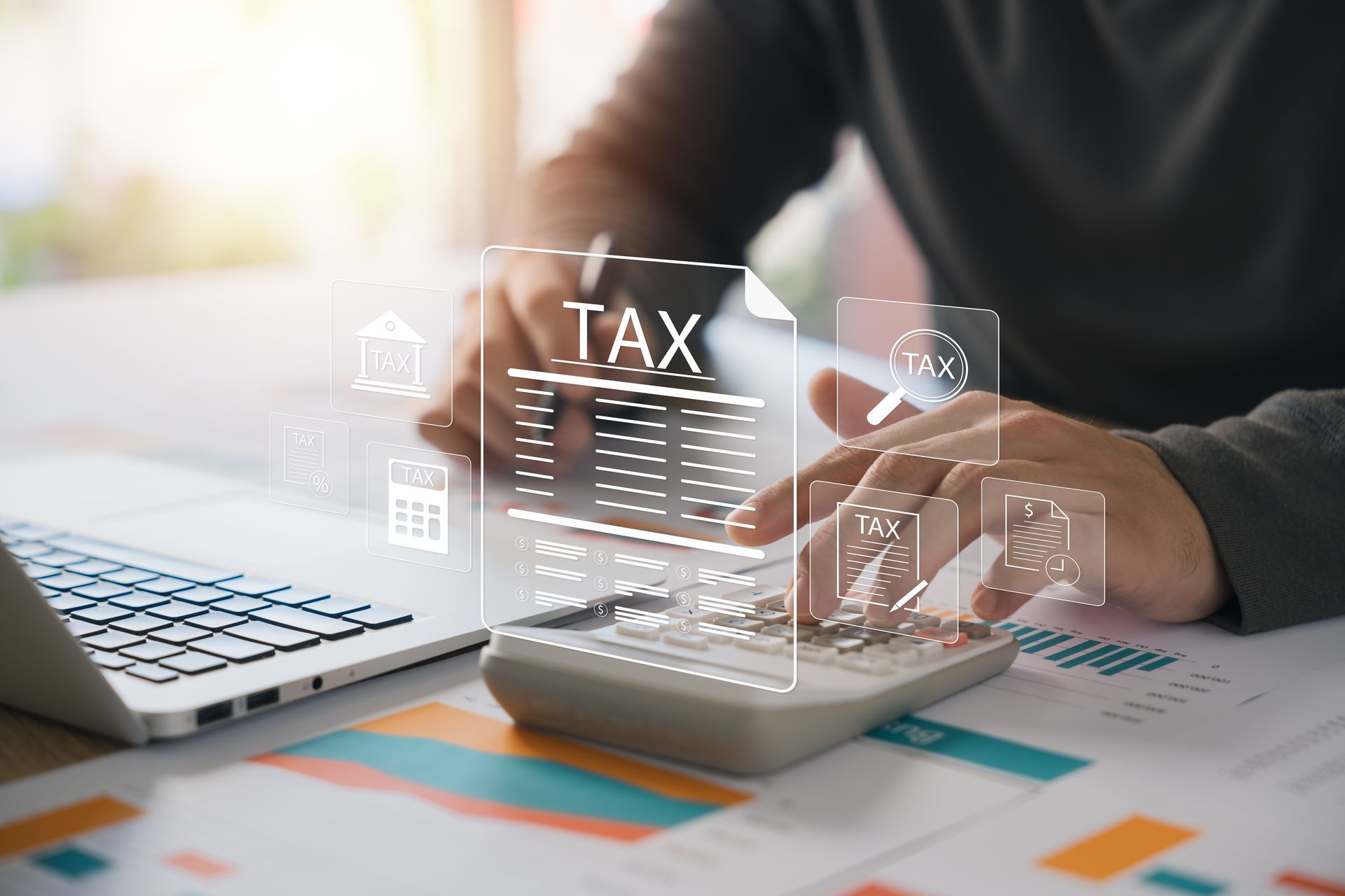 Hands using laptop and calculator with “TAX” hologram over financial charts on a desk