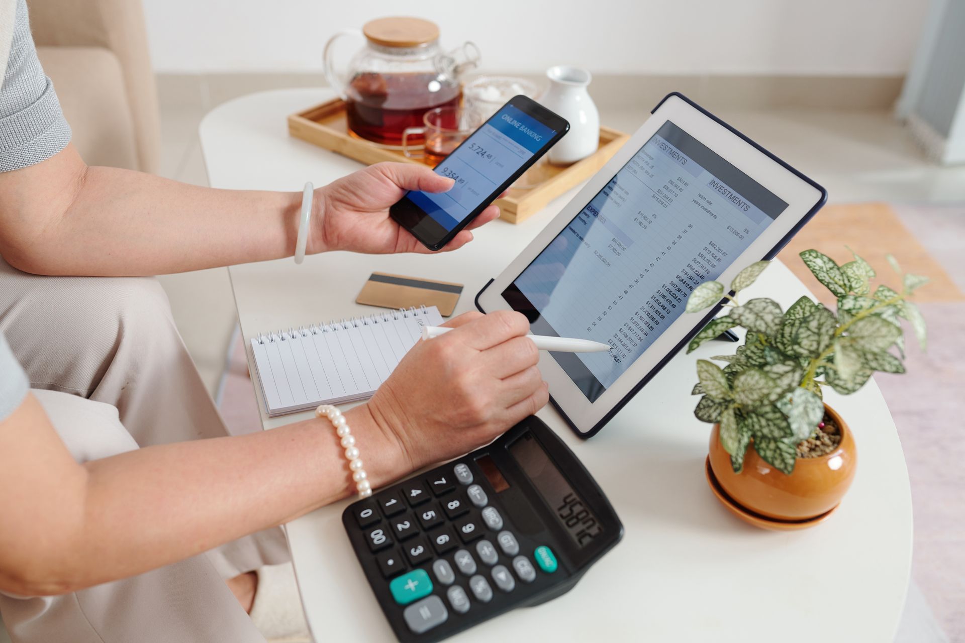 Hands using phone and tablet at a desk with notebooks, coffee, calculator, and a potted plant.