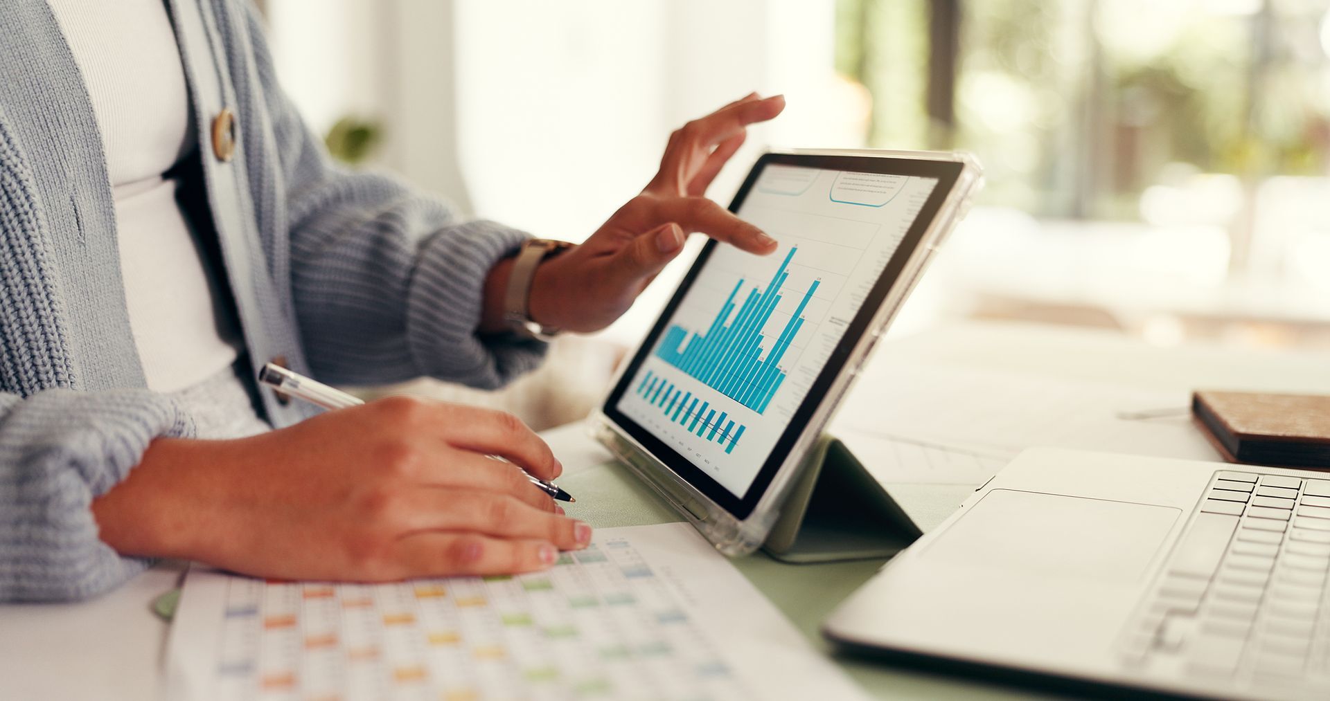 Person using a tablet with a blue bar chart on a desk beside a laptop and keyboard