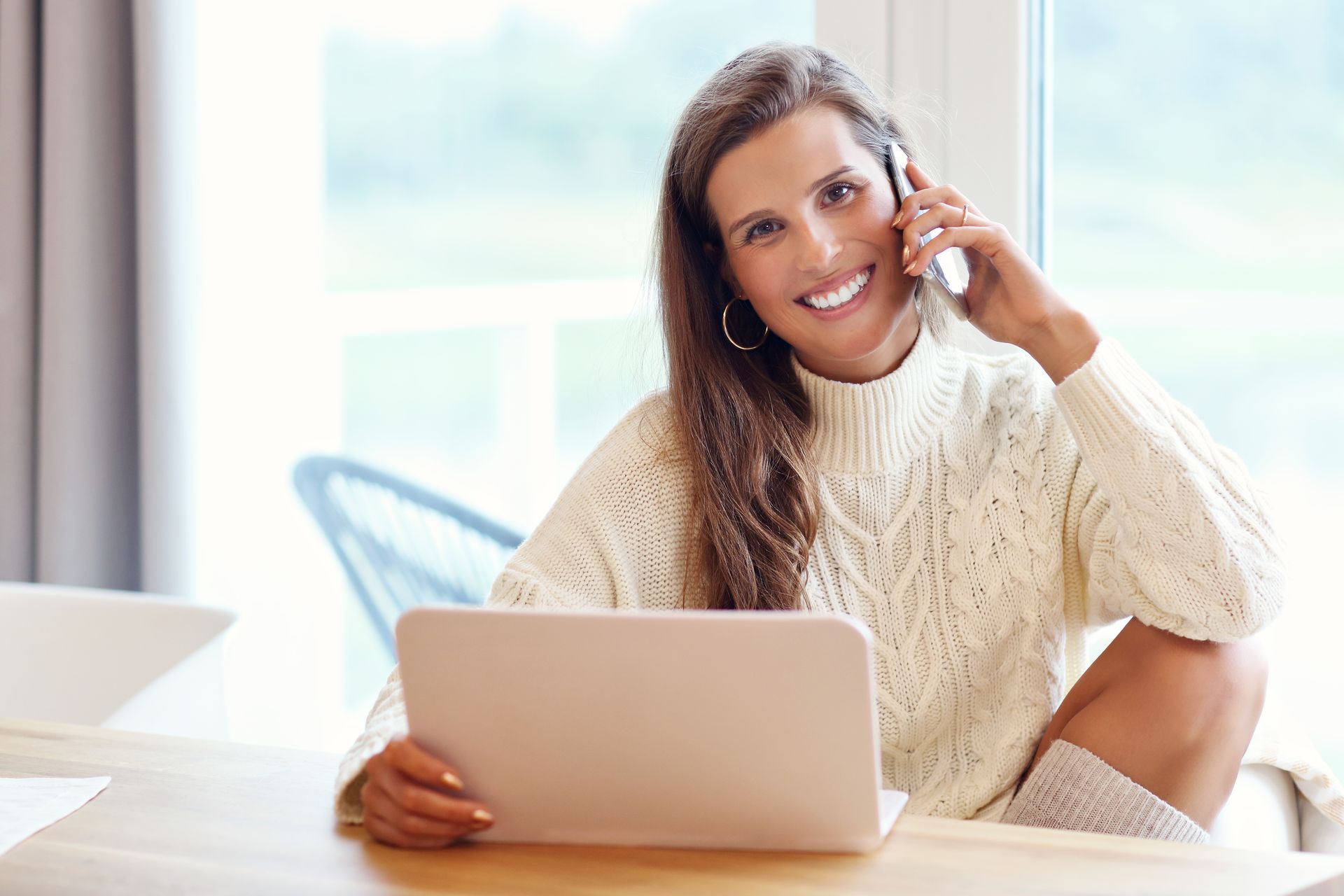 A woman is sitting at a table with a laptop and talking on a cell phone.