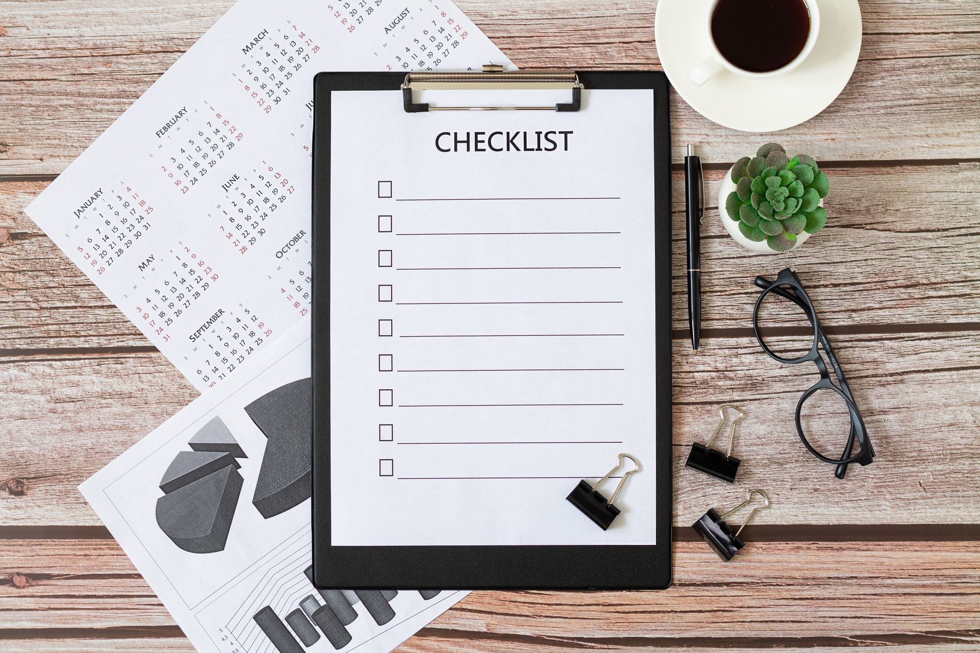 Checklist on a clipboard with documents, a coffee cup, and eyeglasses on a wooden desk.