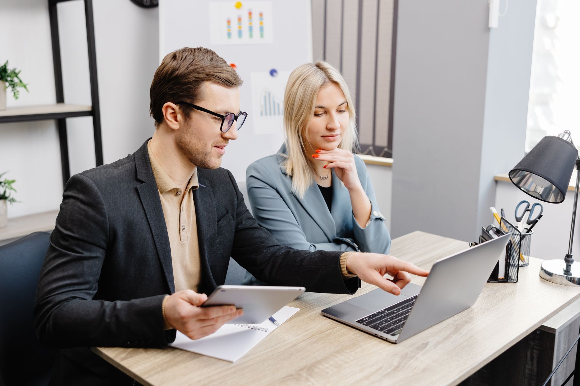 A man and a woman are sitting at a desk looking at a laptop.