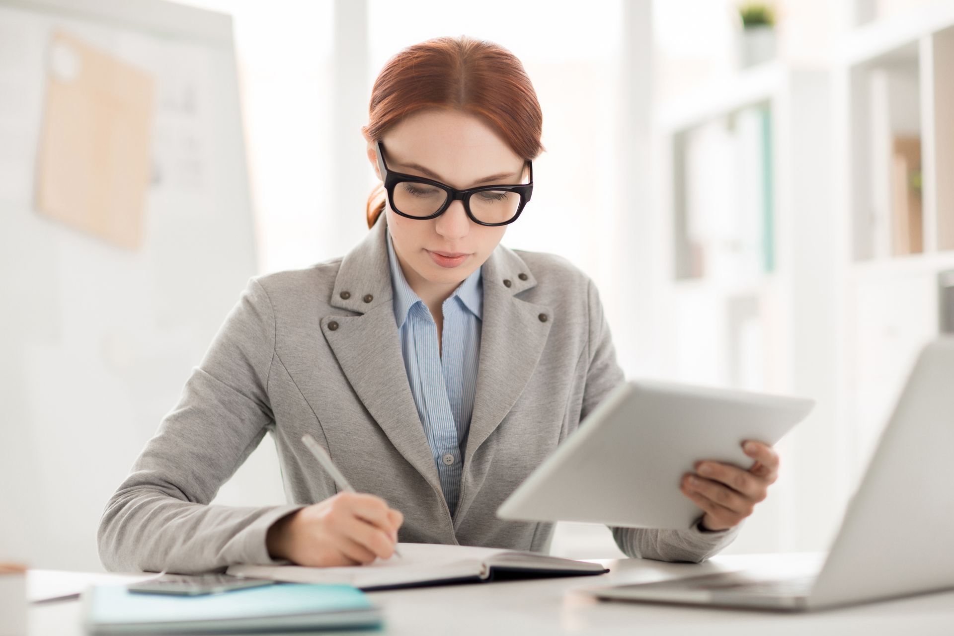 A woman is sitting at a desk with a laptop and a tablet.