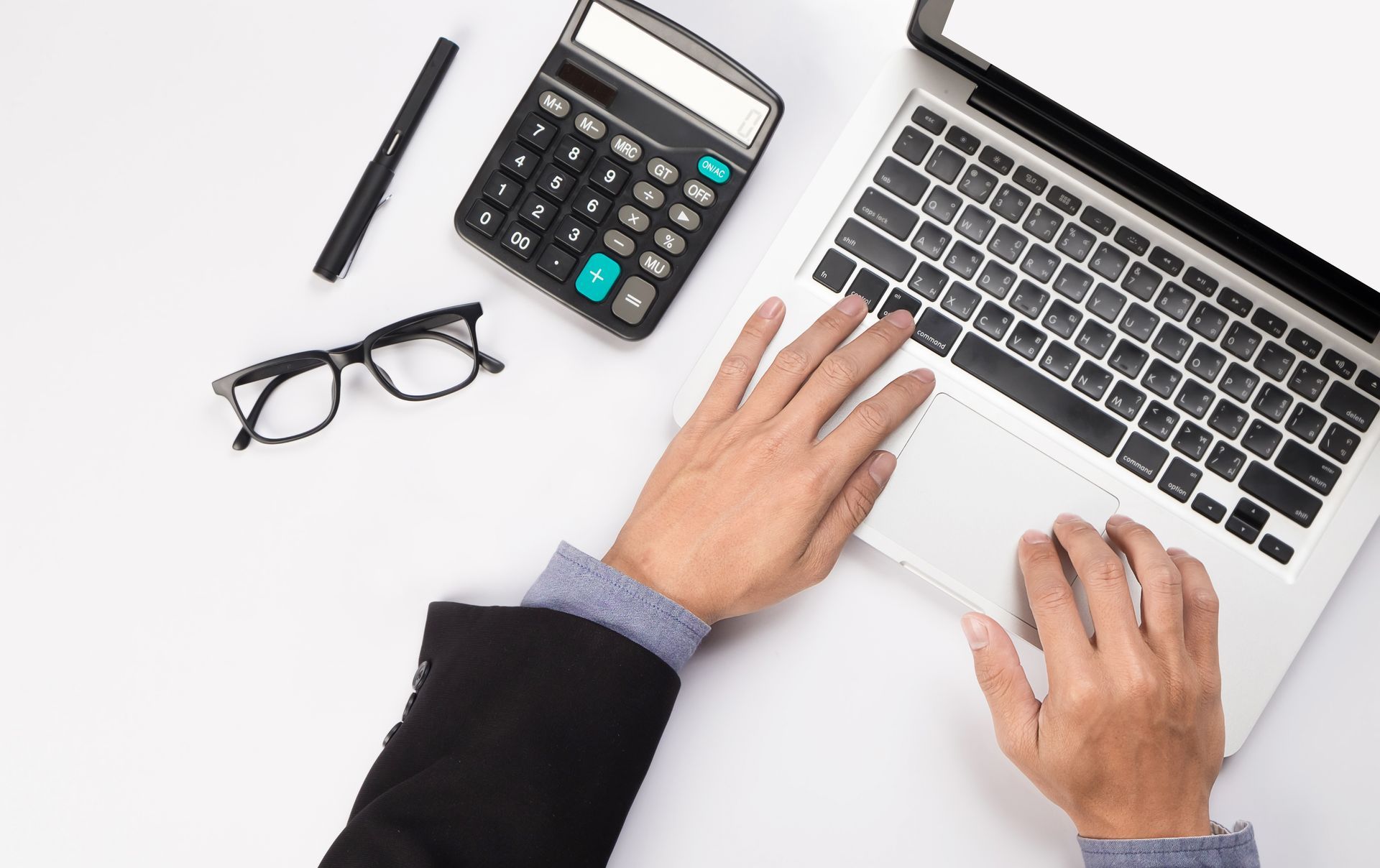 A person is typing on a laptop computer next to a calculator and glasses.
