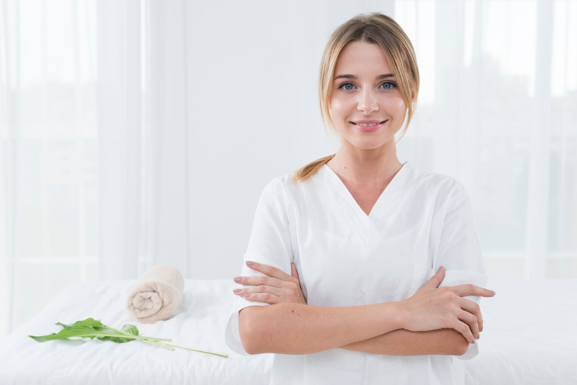 A woman in a white scrub is standing with her arms crossed in front of a massage table.