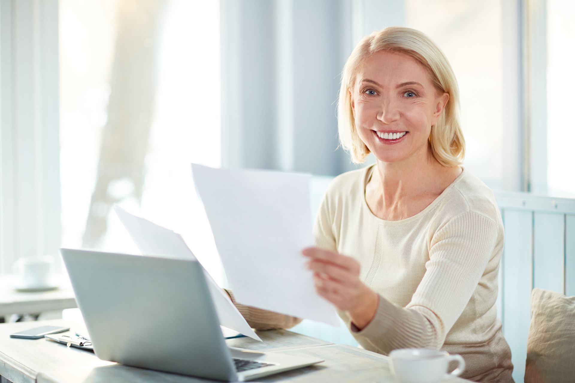 A woman is sitting at a desk with a laptop and holding a piece of paper.