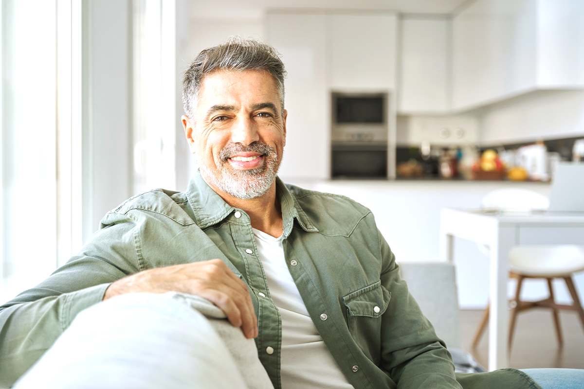 Homem sorri, sentado no sofá em uma sala iluminada. Ele veste uma camisa verde.