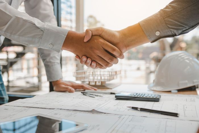 Two men are shaking hands over a table with blueprints.