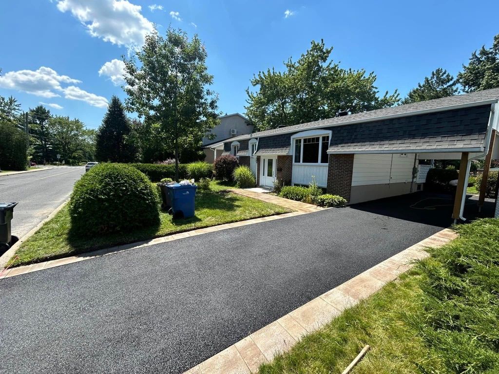 A black asphalt driveway leads to a suburban house with a carport on a sunny day.