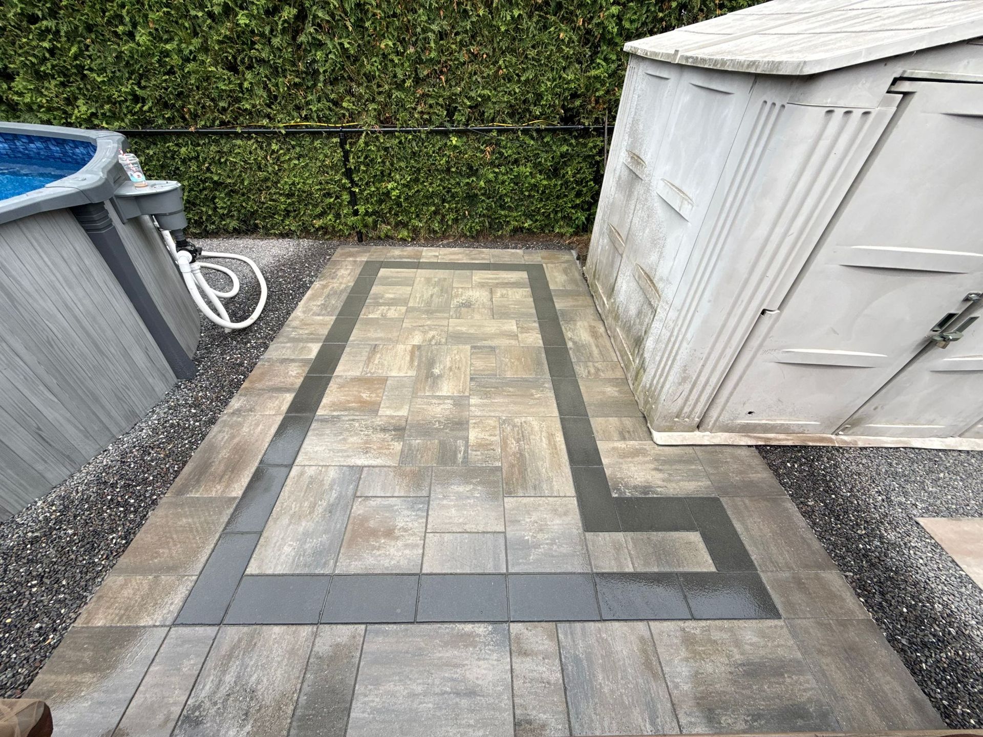 Patio with gray stone pavers, dark border, near pool and shed, surrounded by pebbles and a green hedge.