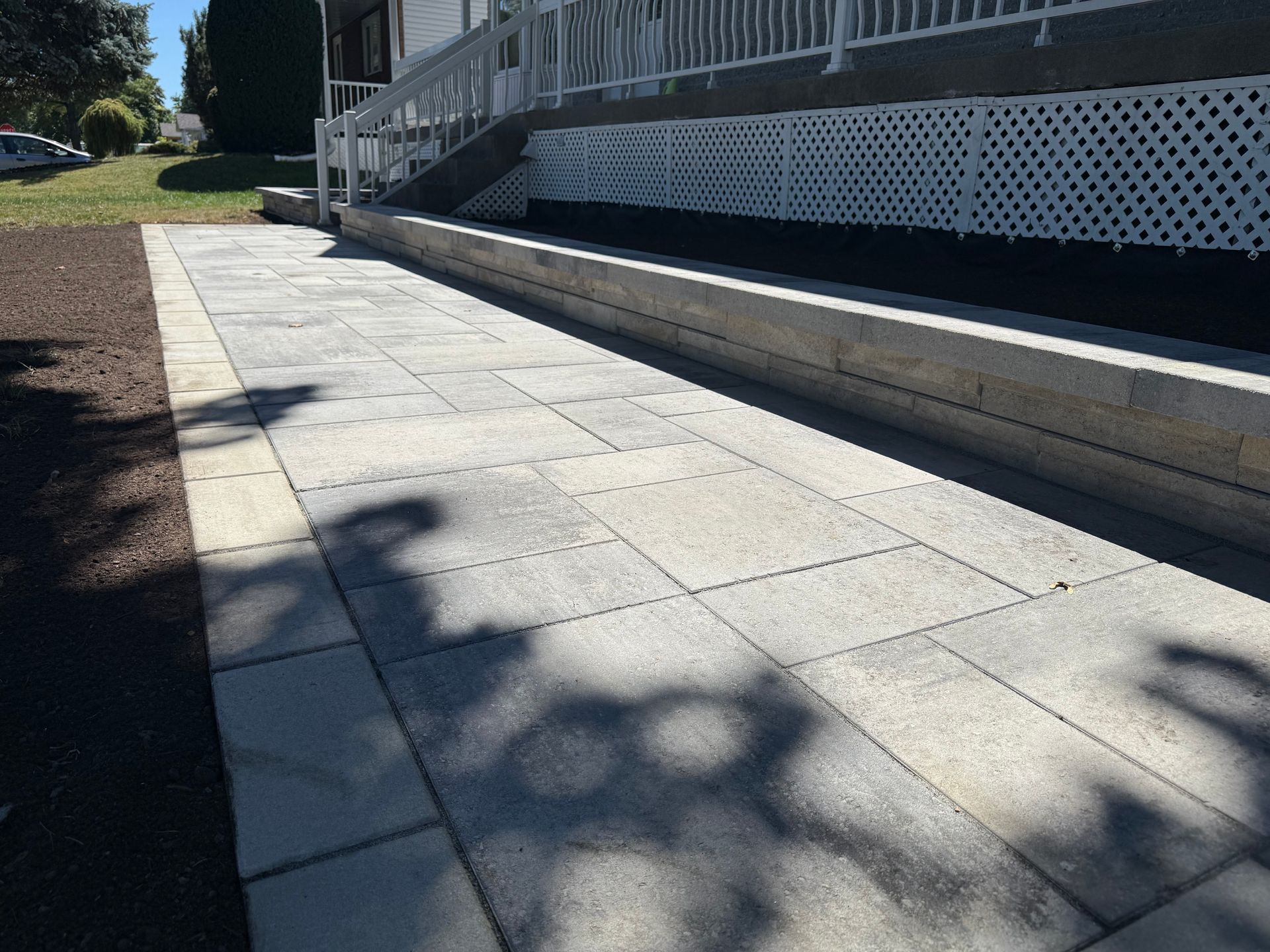 Gray paver walkway alongside a building with white railing and lattice. Shadows cast by trees.