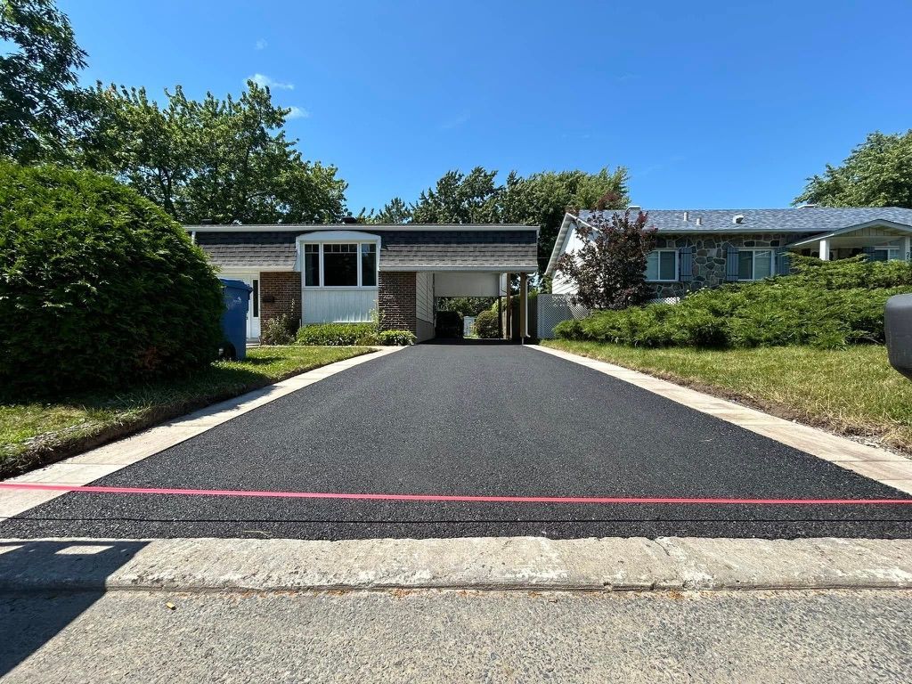 Newly paved black asphalt driveway leading to a house, with a blue sky and green trees.
