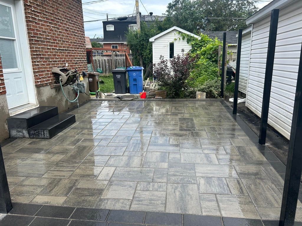 Newly paved patio with gray pavers; brick wall, sheds, and trees in background.