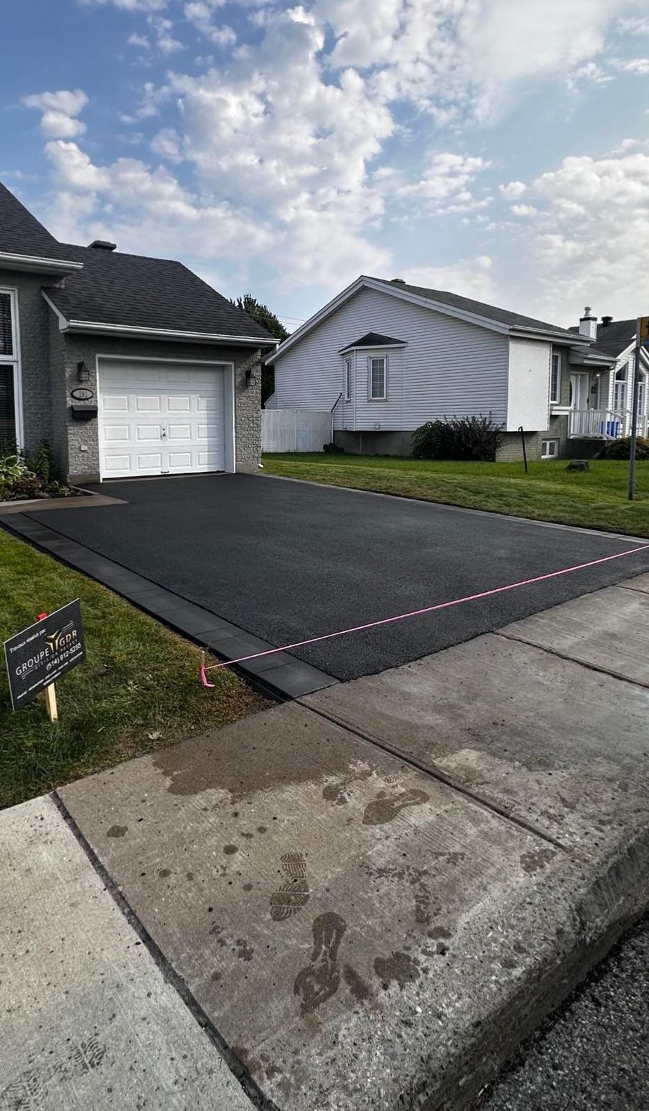 Newly paved black asphalt driveway next to a grass lawn and sidewalk, with a white garage door in the background.