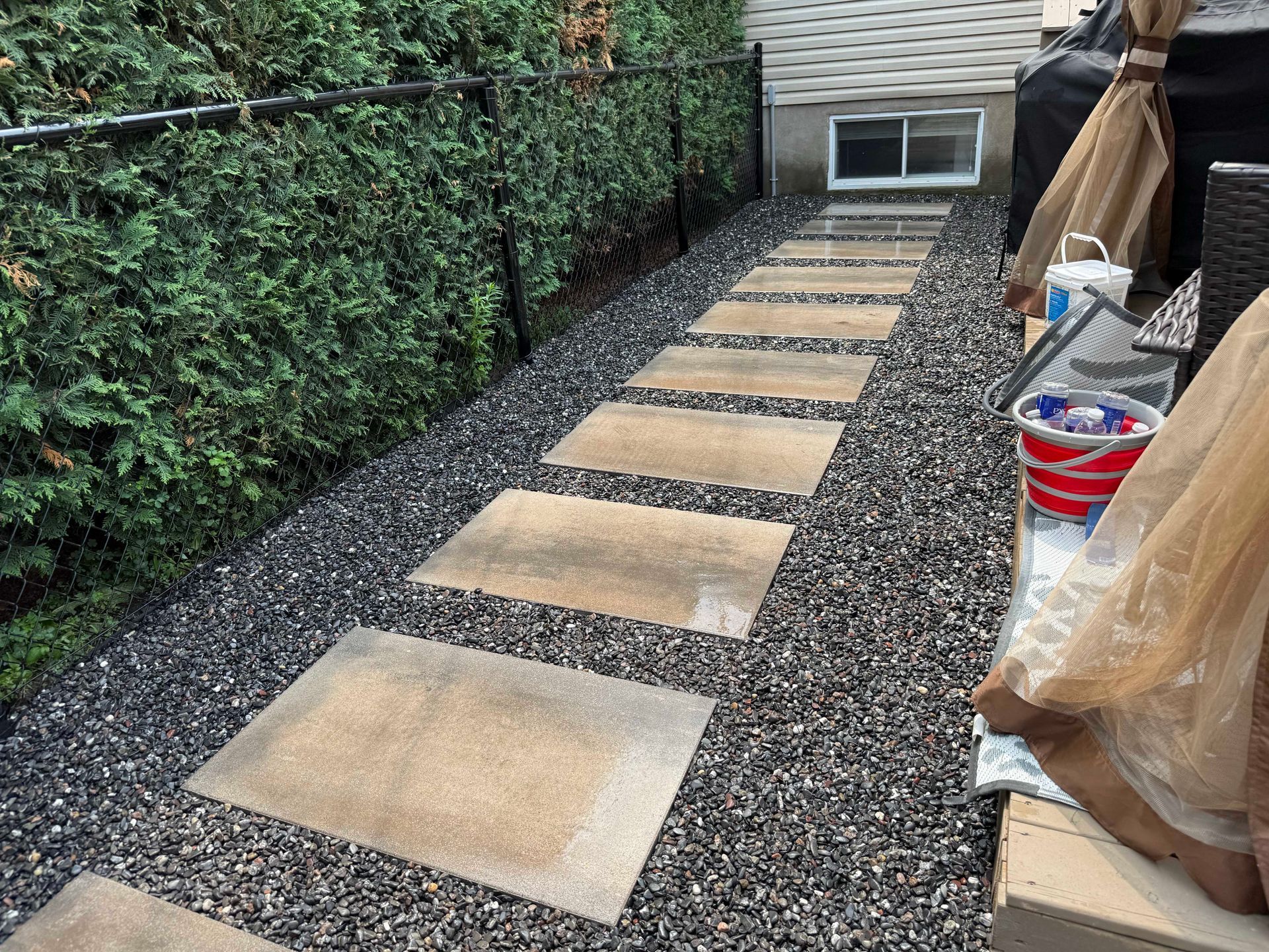 Stone pathway through gravel next to a hedge and fence.