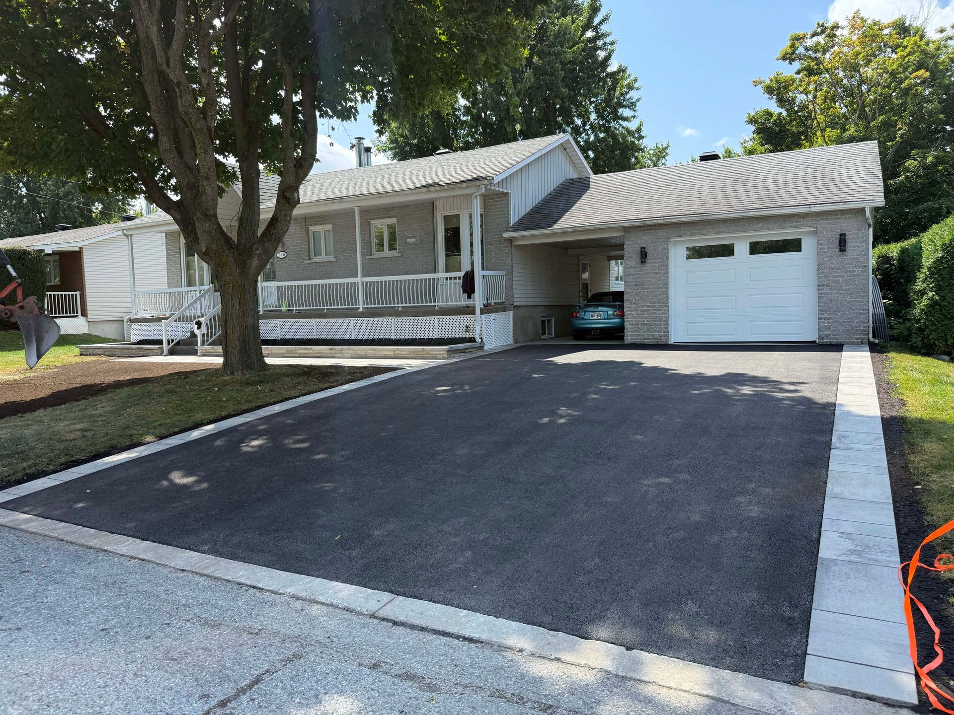 Gray house with a driveway. Carport on the right, porch on the left. Dark asphalt driveway with stone border.