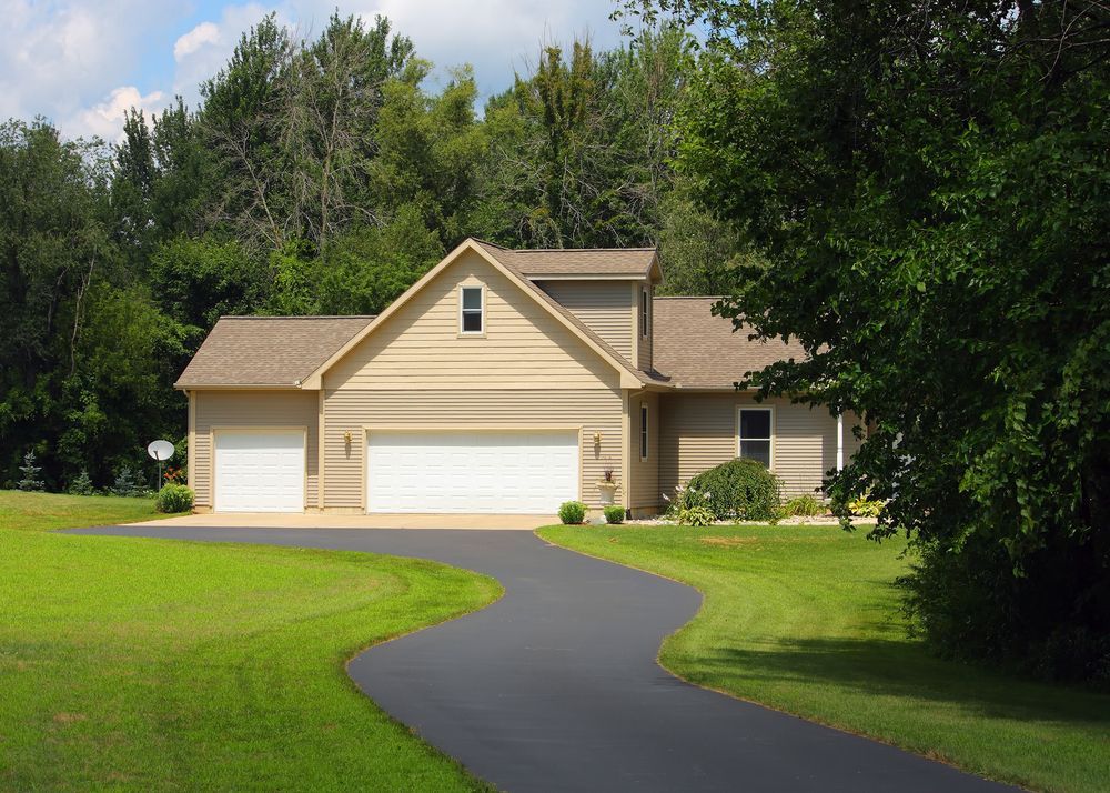 House with a three-car garage, beige siding, and a winding black driveway surrounded by green grass and trees.