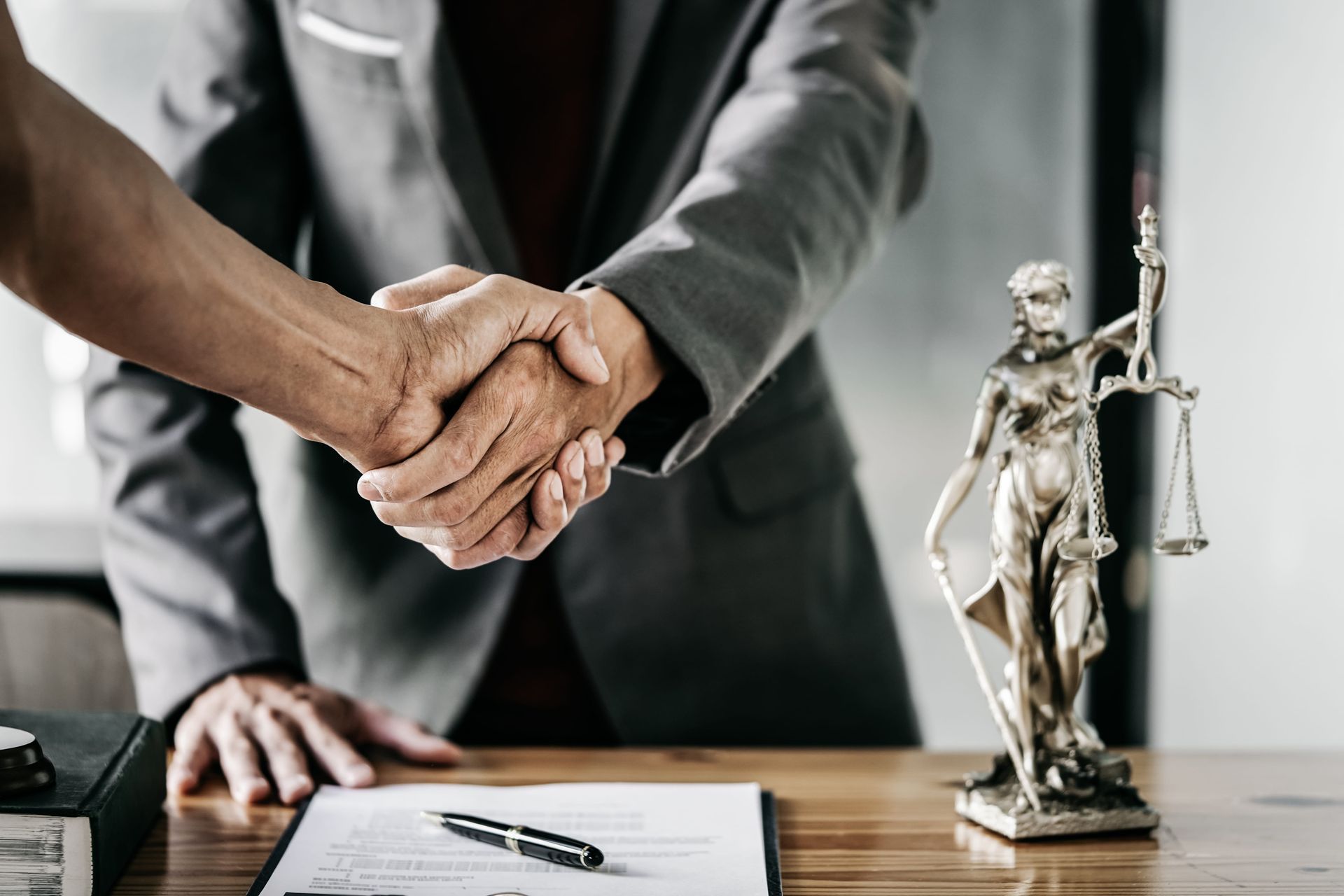 Two people shaking hands across a desk beside a legal statue and signed document