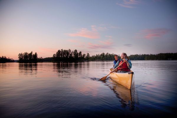 Two people canoeing on a calm lake at sunset, with a tree-lined shore in the distance.