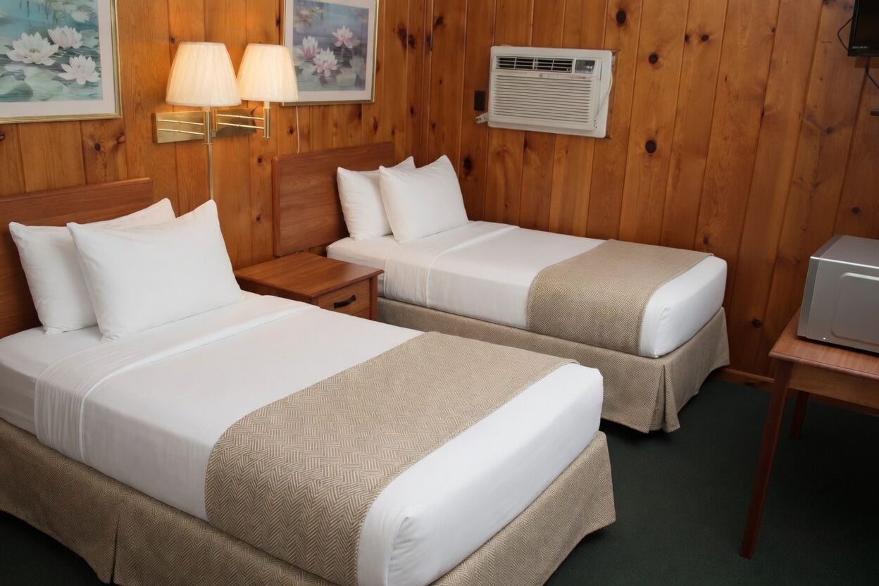 Small motel room with two white beds, wood-paneled walls, lamps, and a desk