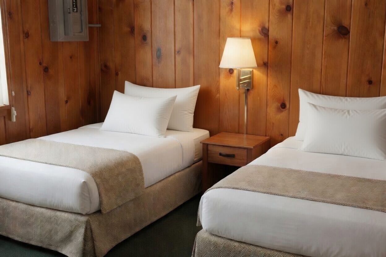 Two twin beds with white bedding in a wood-paneled room, lit by a bedside lamp.