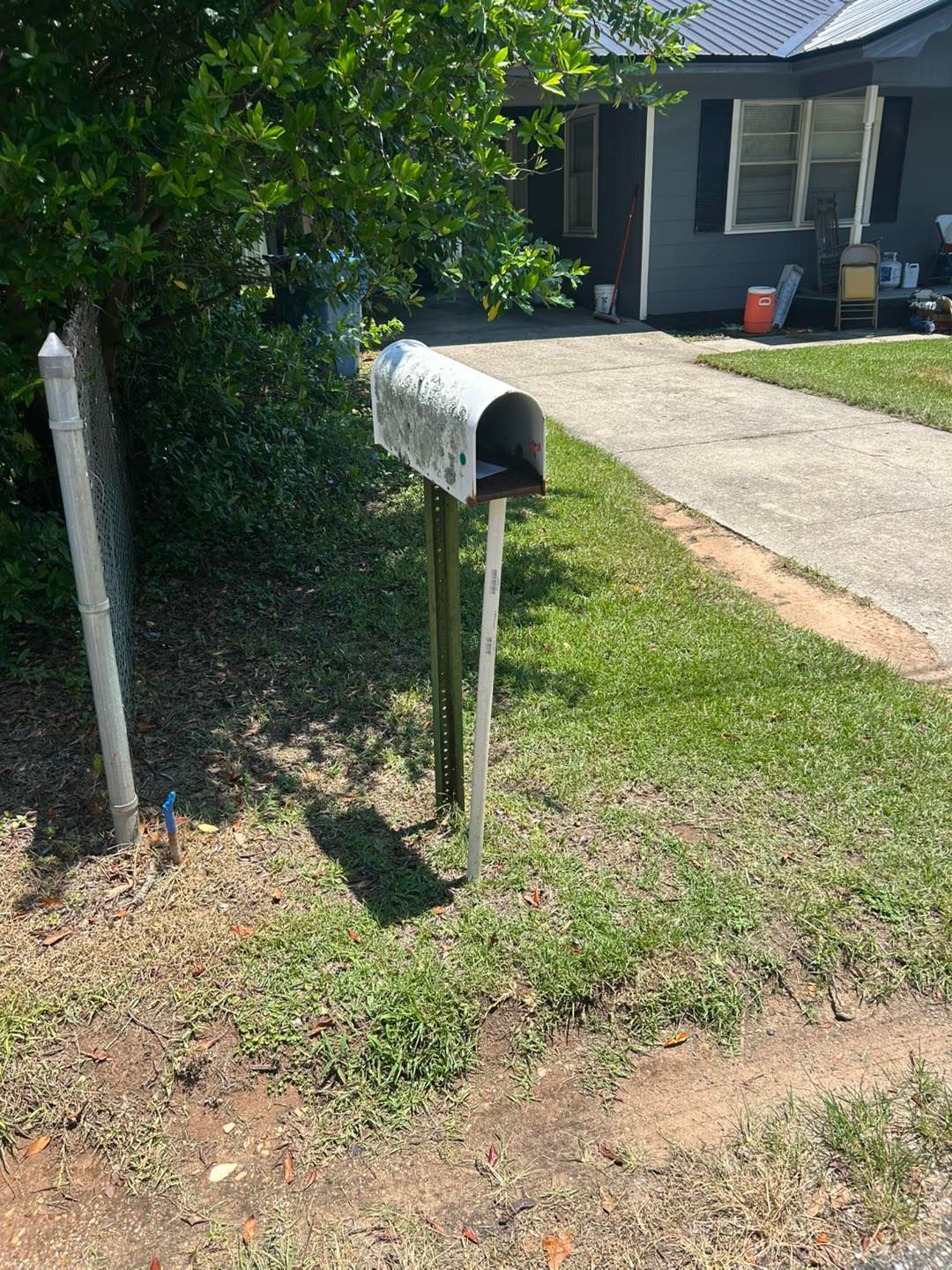 Weathered metal mailbox on a wooden post beside a driveway and a house with blue siding.