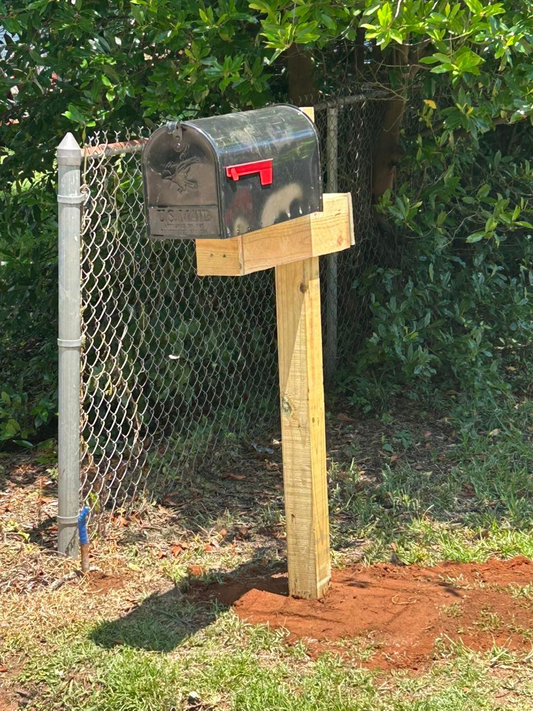 Black mailbox on a wooden post next to a chain-link fence, set in red-brown soil and grass.