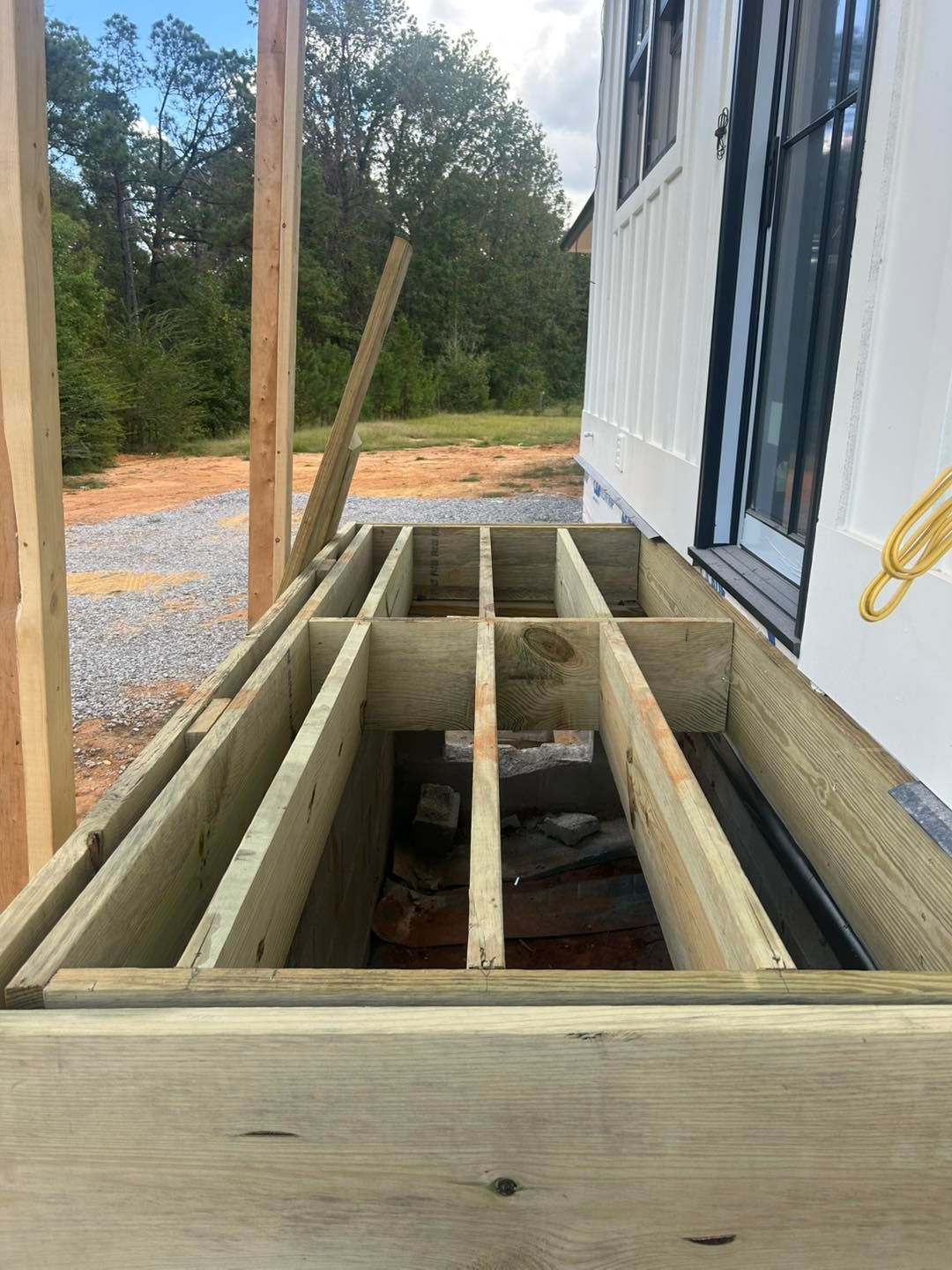 Wooden deck frame under construction next to a white house with window. Gravel ground.