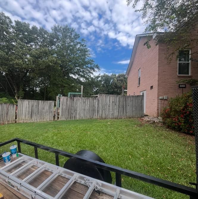 Backyard with weathered wooden fence, pink house, green grass, and a trailer with ladder and paint cans.