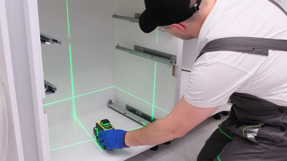 A person uses a laser level to install drawer slides inside a white cabinet.