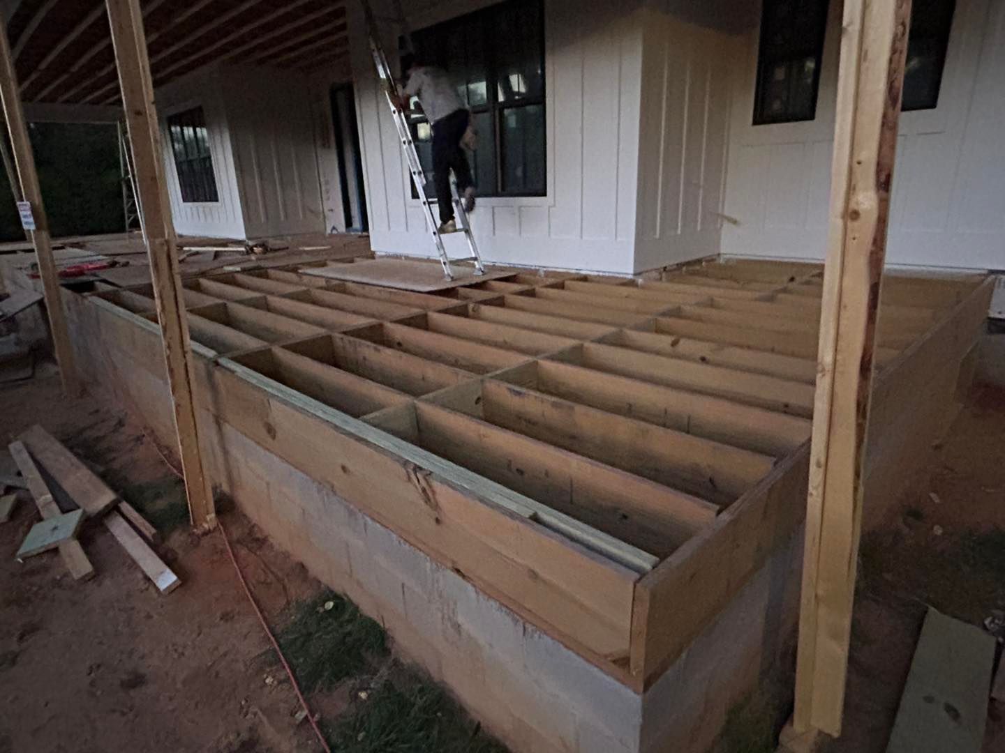 Construction of a wooden deck. A person climbs a ladder near a house with white siding.