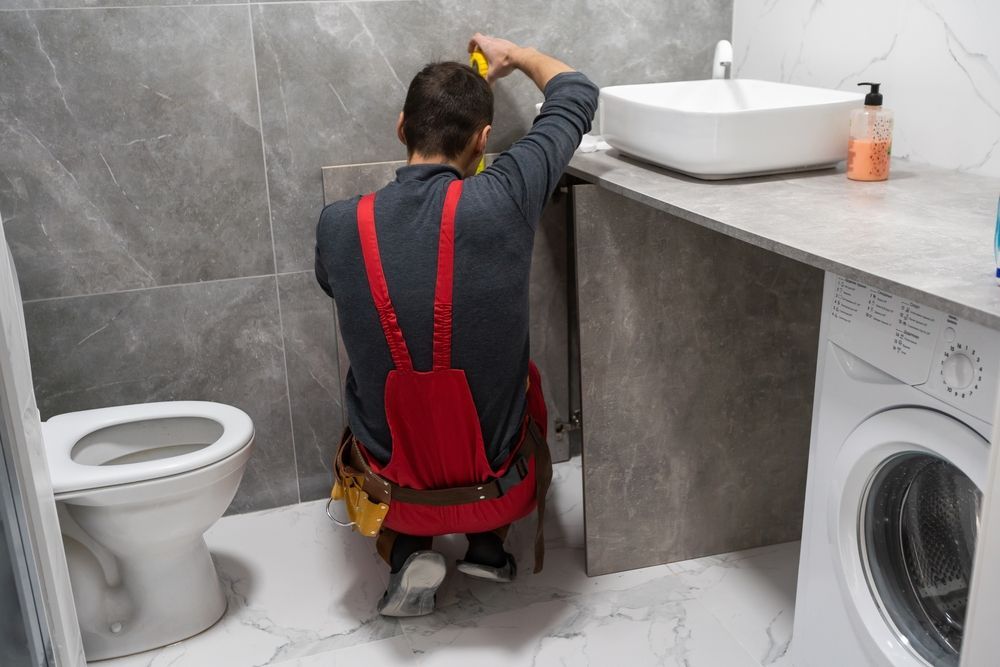 Plumber installing a fixture in a gray bathroom, near a toilet, sink, and washing machine.