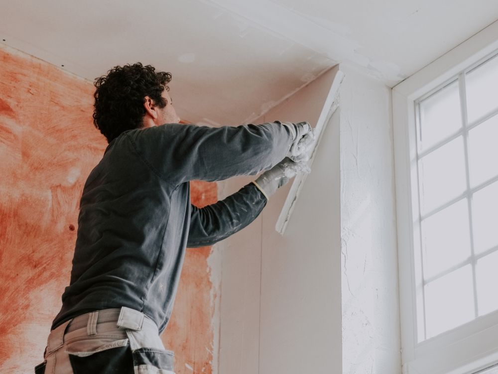 Man using a trowel to apply plaster to a wall. Room has an orange wall, white trim, and a window.