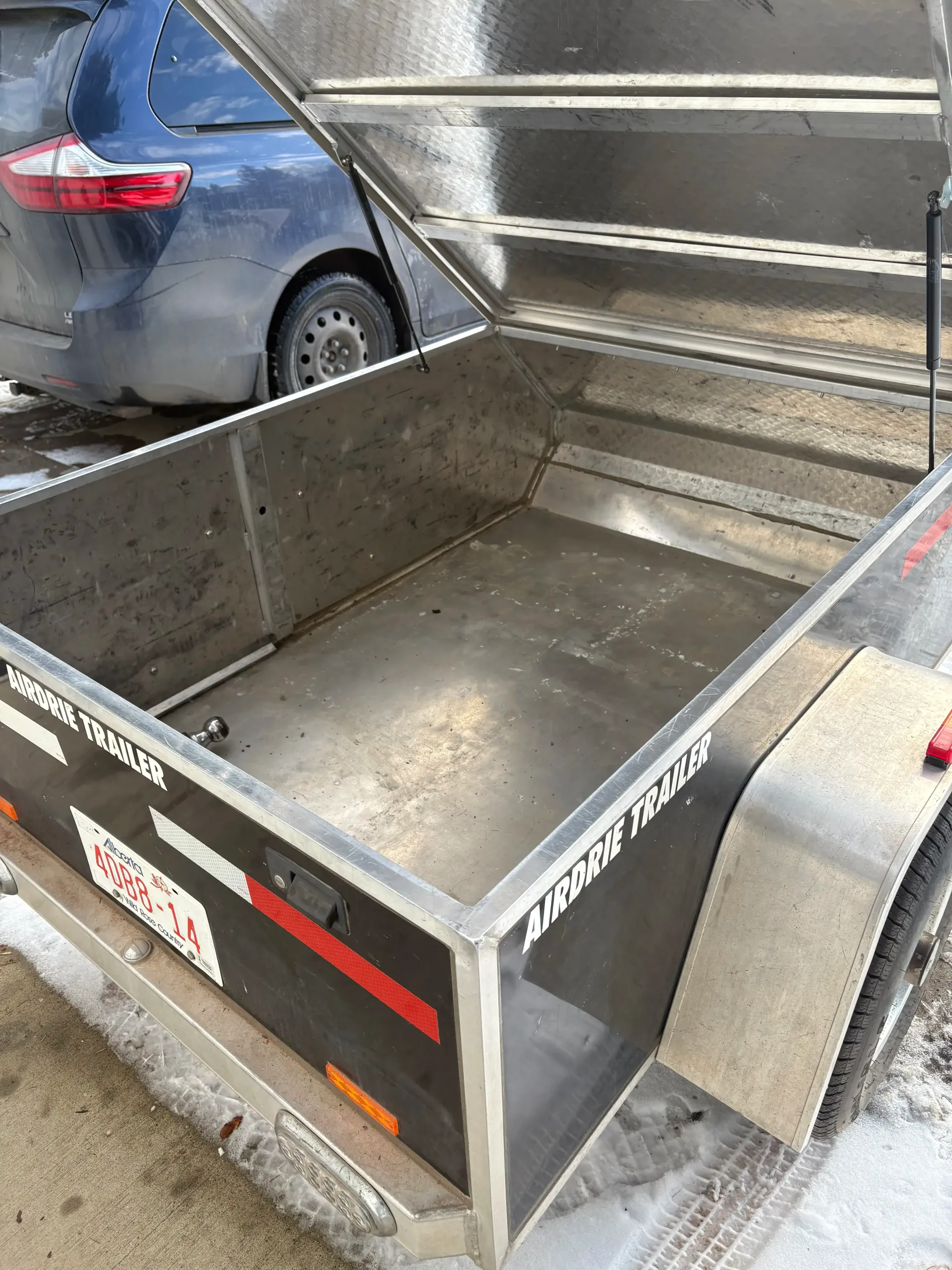 Open aluminum utility trailer with a black exterior; car is in the background.