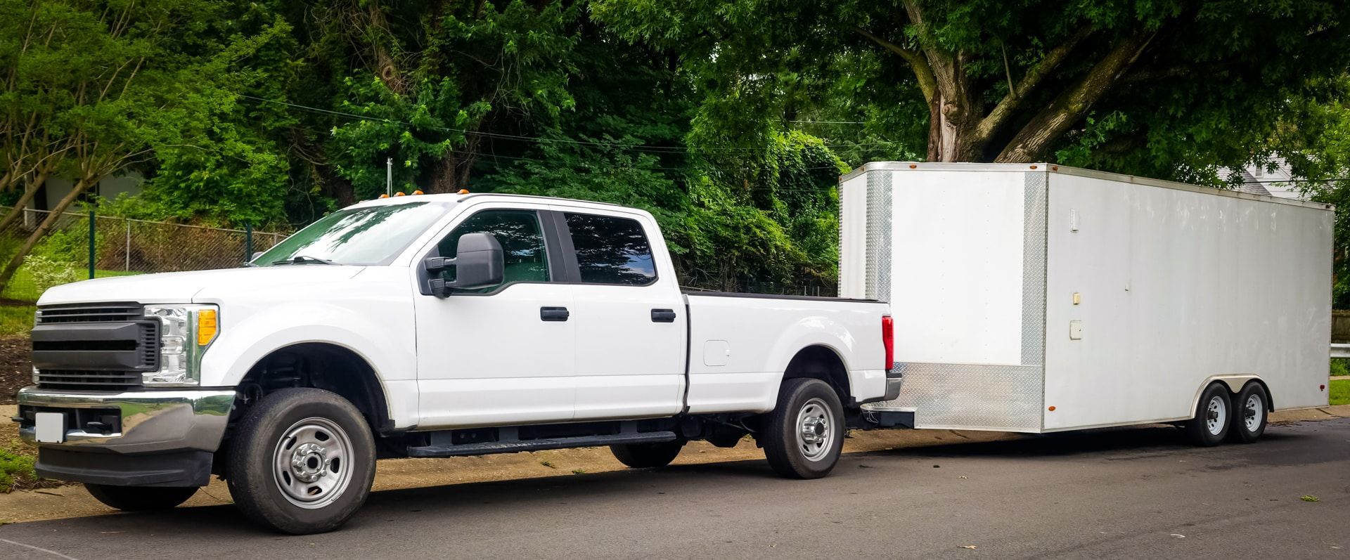 White pickup truck towing a large white trailer on a road, trees in the background.