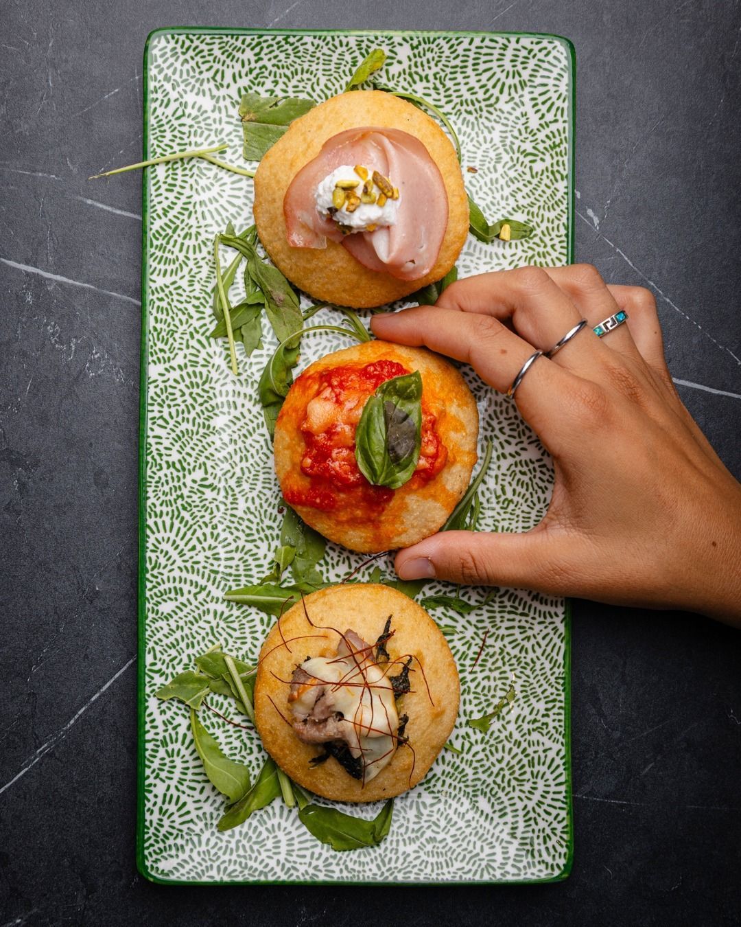 Tre antipasti rotondi di pane piatto conditi con prosciutto, salsa di pomodoro e funghi su un piatto verde. Ne tengo uno in mano.