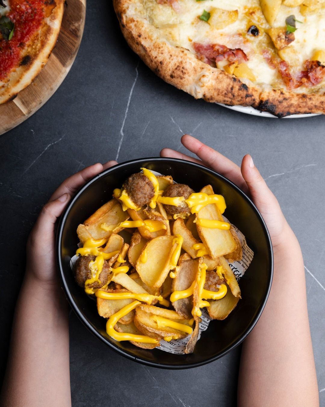 Mani che tengono una ciotola di spicchi di patate con salsa al formaggio, con due pizze sullo sfondo su una superficie scura.