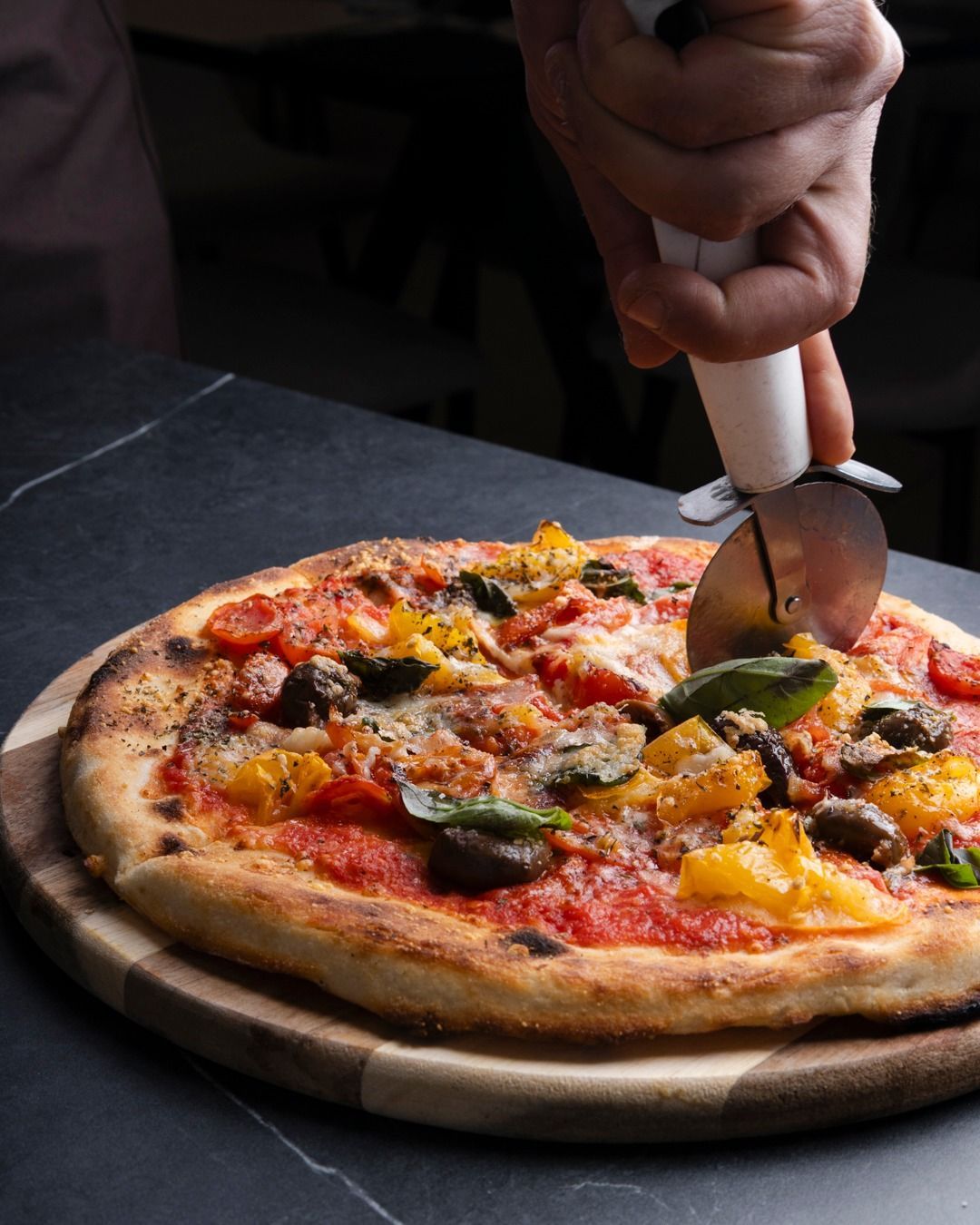 Tagliare a mano una pizza con peperoni gialli, pomodori, basilico e funghi su una tavola di legno.
