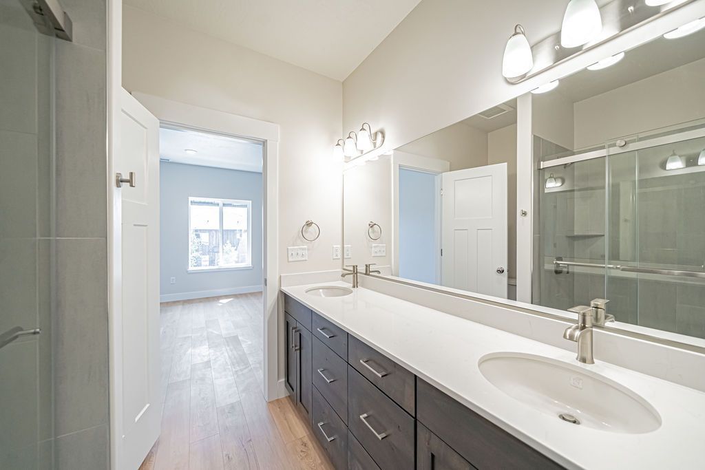 Bathroom with double vanity, large mirror, and open doorway to a bedroom with light wood flooring.