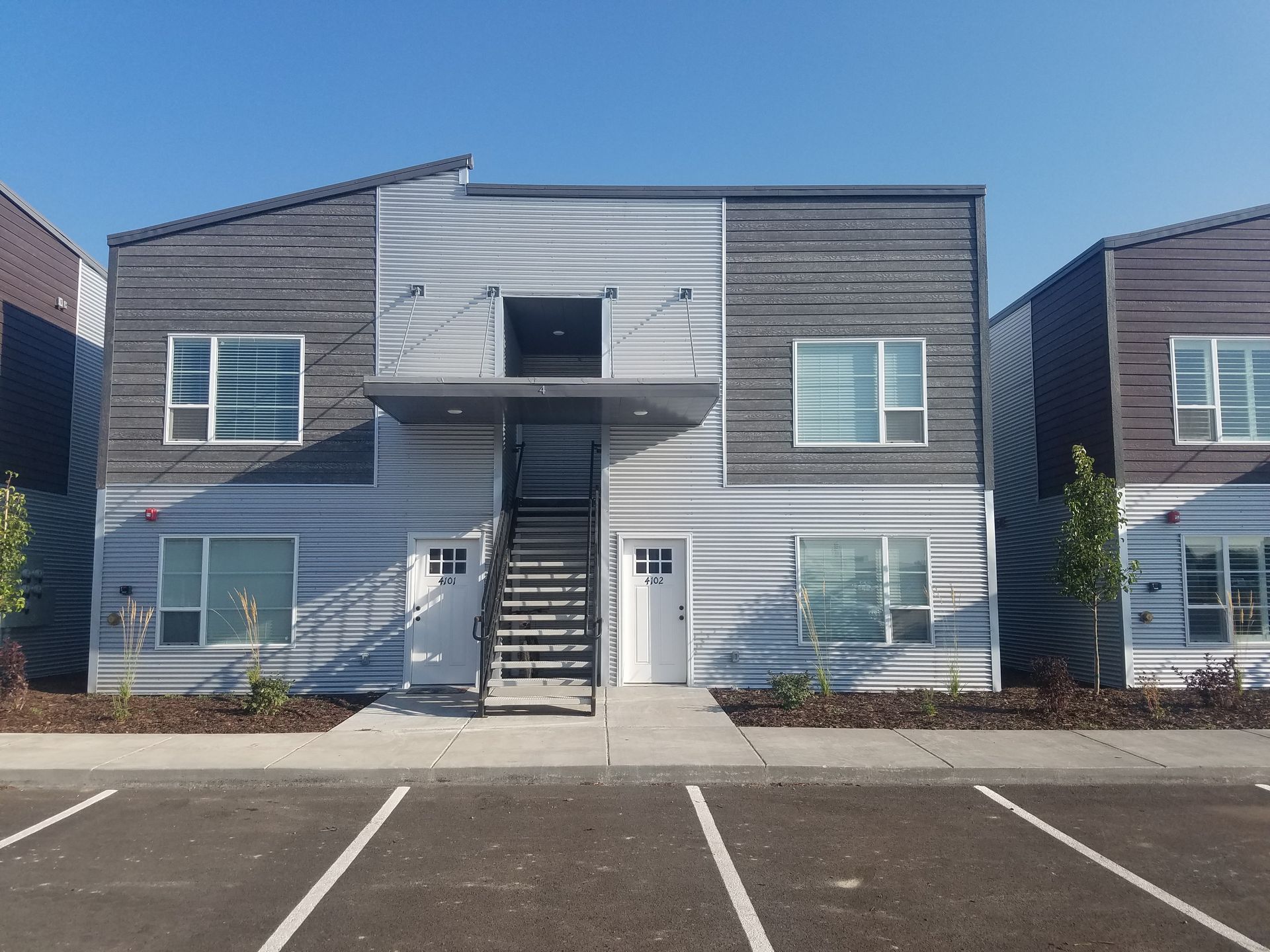 Two-story modern apartment building with metal siding, stairs leading to a second-floor entrance, blue sky background.