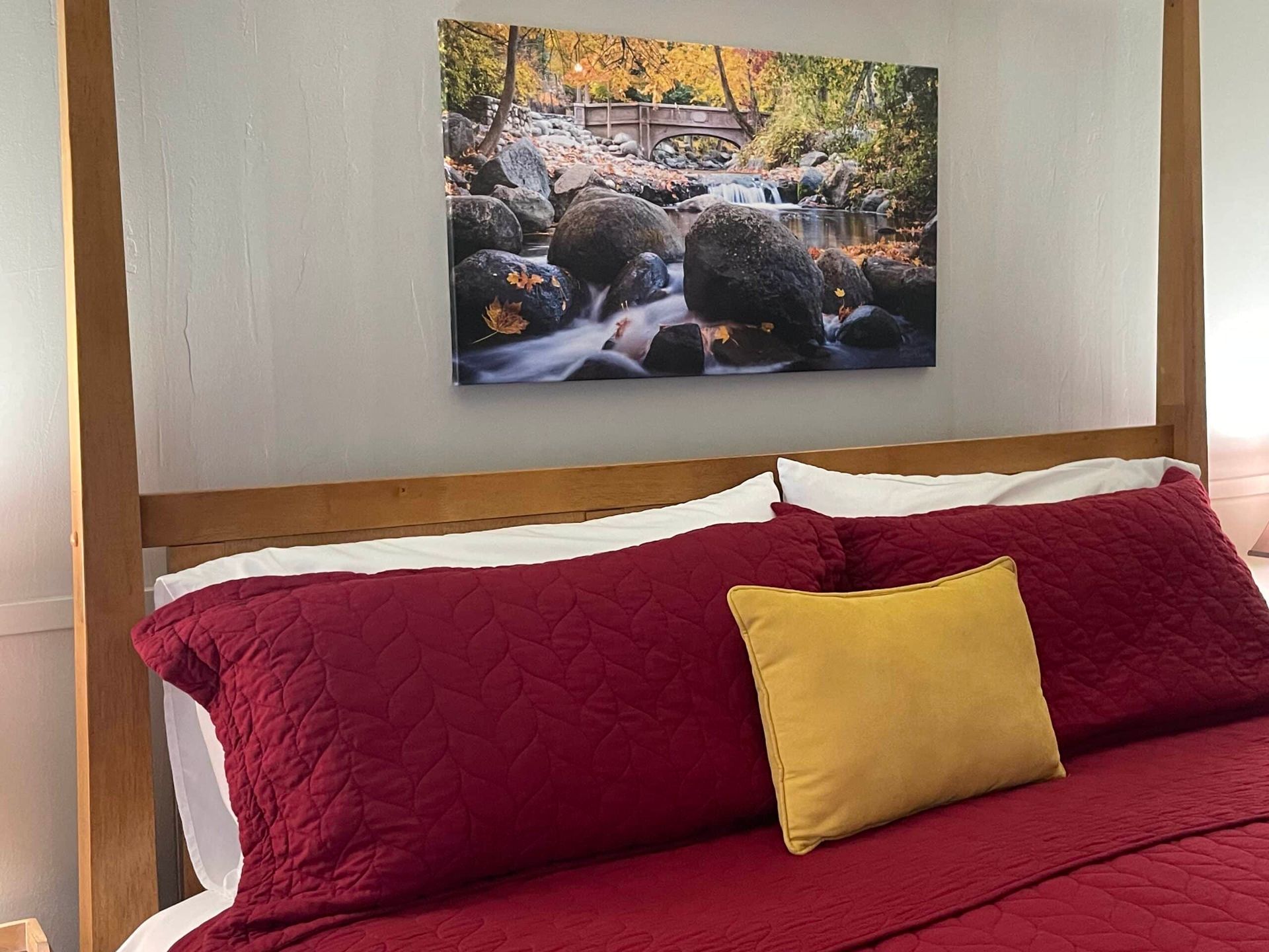 A bed with red sheets and pillows and a painting on the wall above it.