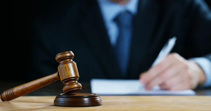 Gavel on a wooden desk with a person in a suit writing in the background.