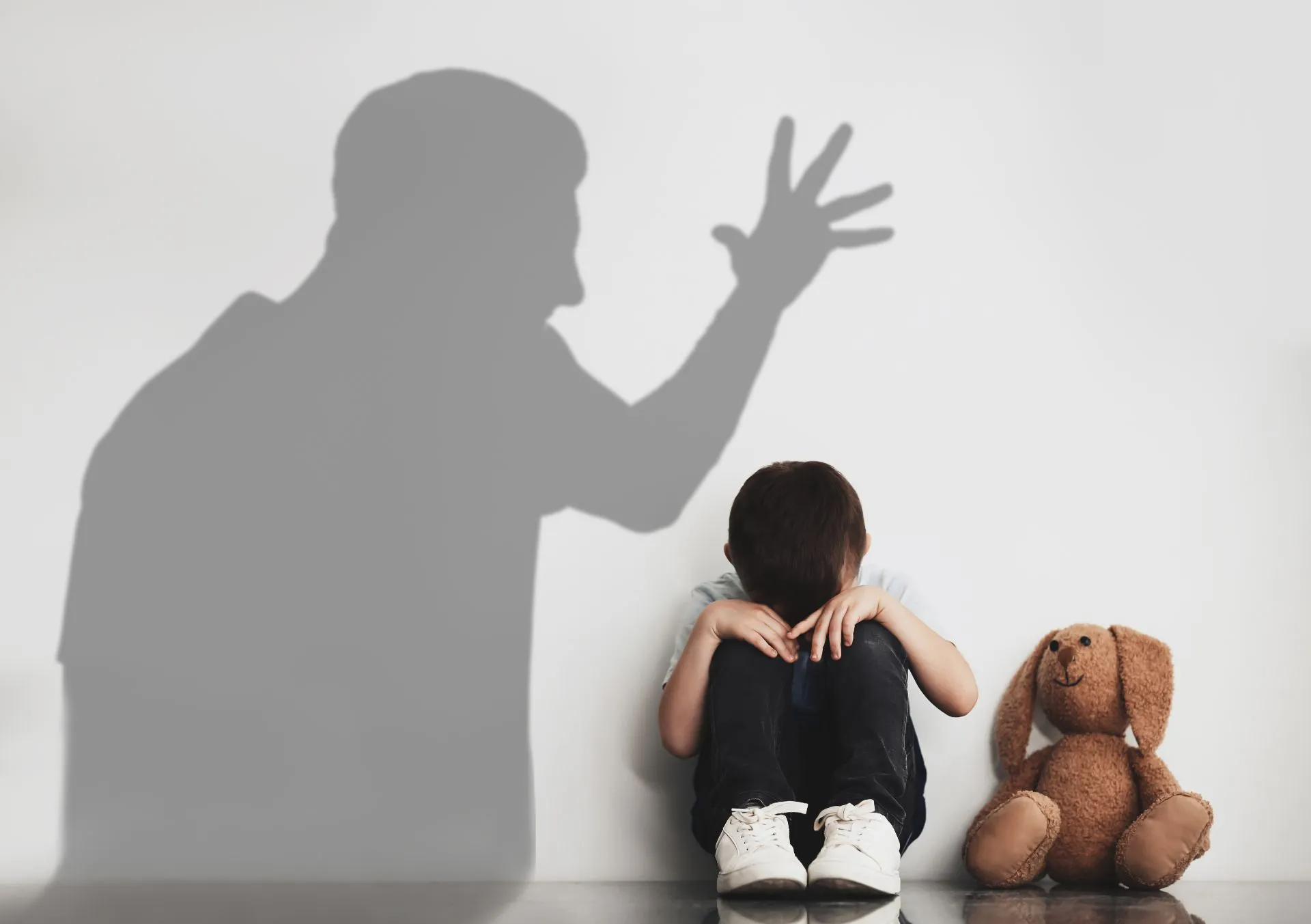 Child in distress, hiding from shadow of a yelling adult; teddy bear nearby.