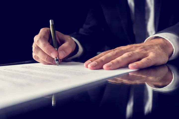 Person in a suit signing a document with a pen on a dark, reflective surface.