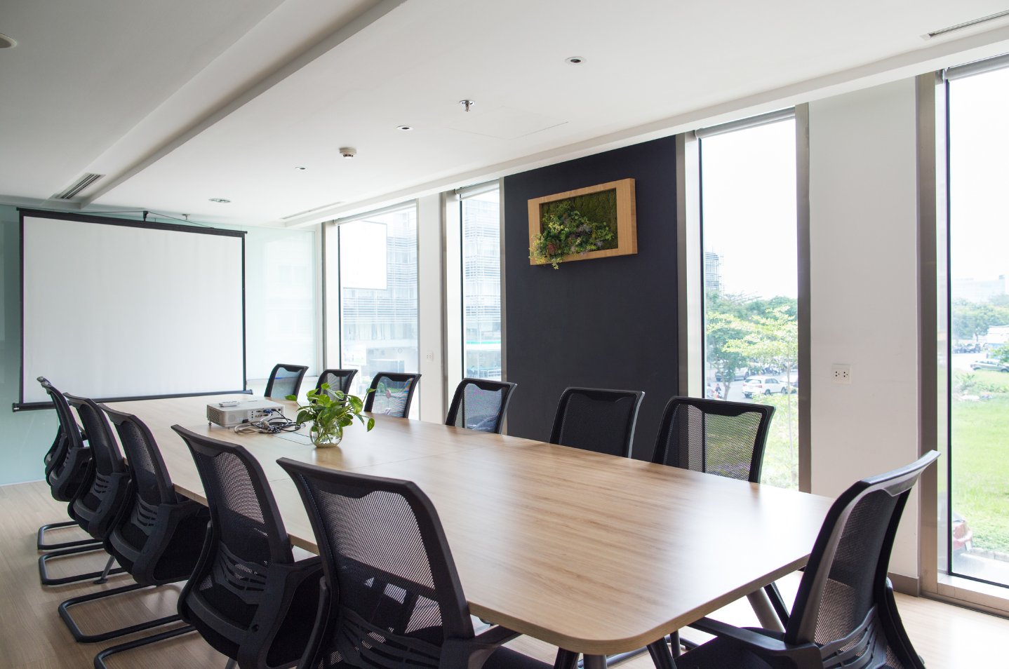 Modern conference room with long wooden table, black chairs, projector screen, and large windows.