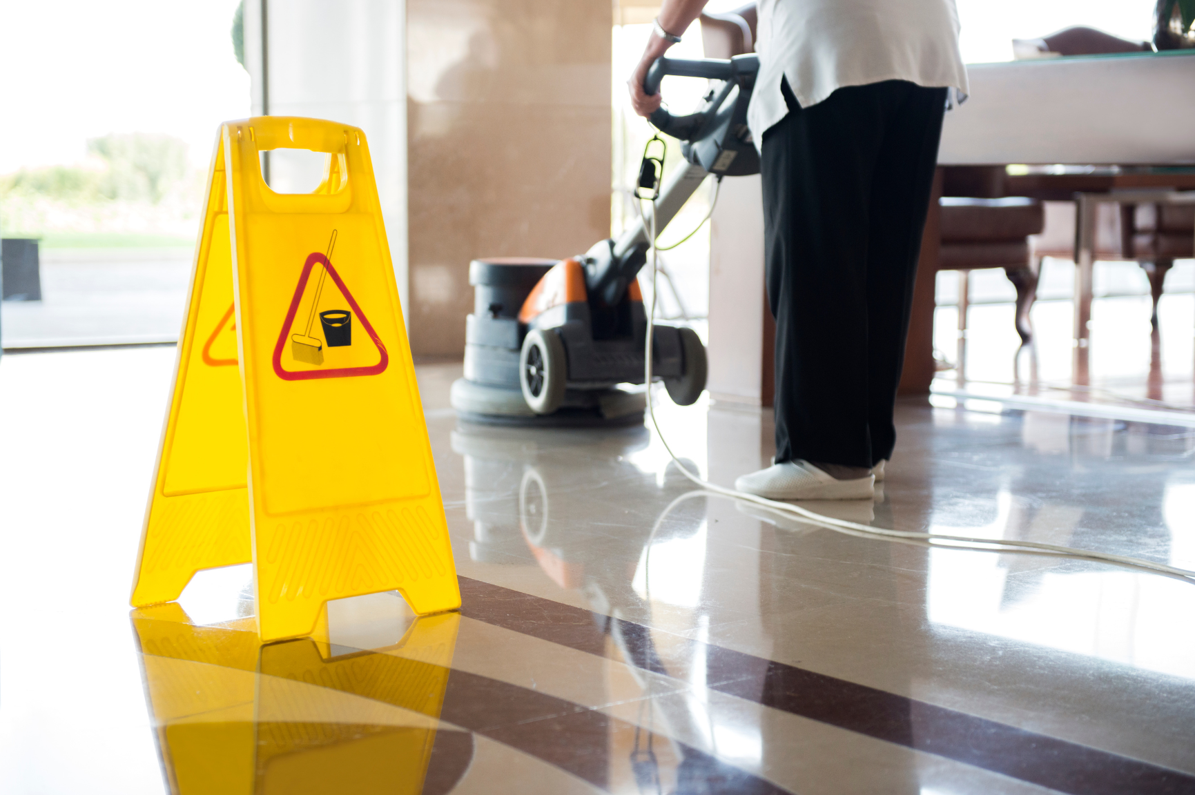Yellow wet floor sign on polished floor, person cleaning with a floor buffer.