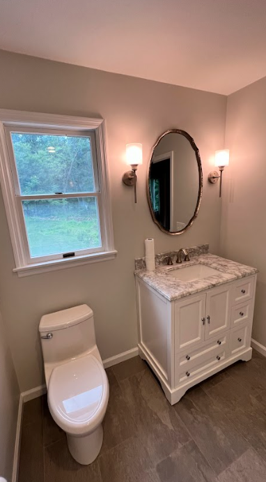 A bathroom with a toilet , sink , mirror and window.