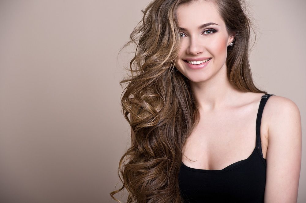 Woman with long, wavy brown hair smiles, wearing a black tank top against a neutral background.