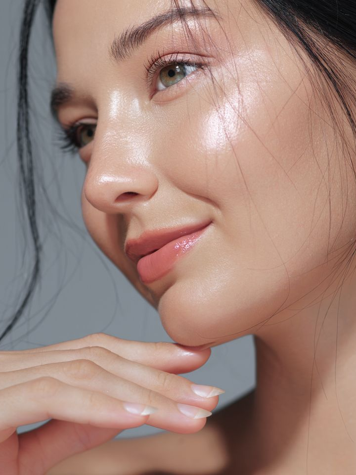 Close-up of a woman’s face with smooth, glowing skin and soft pink lips, resting her chin on her hand